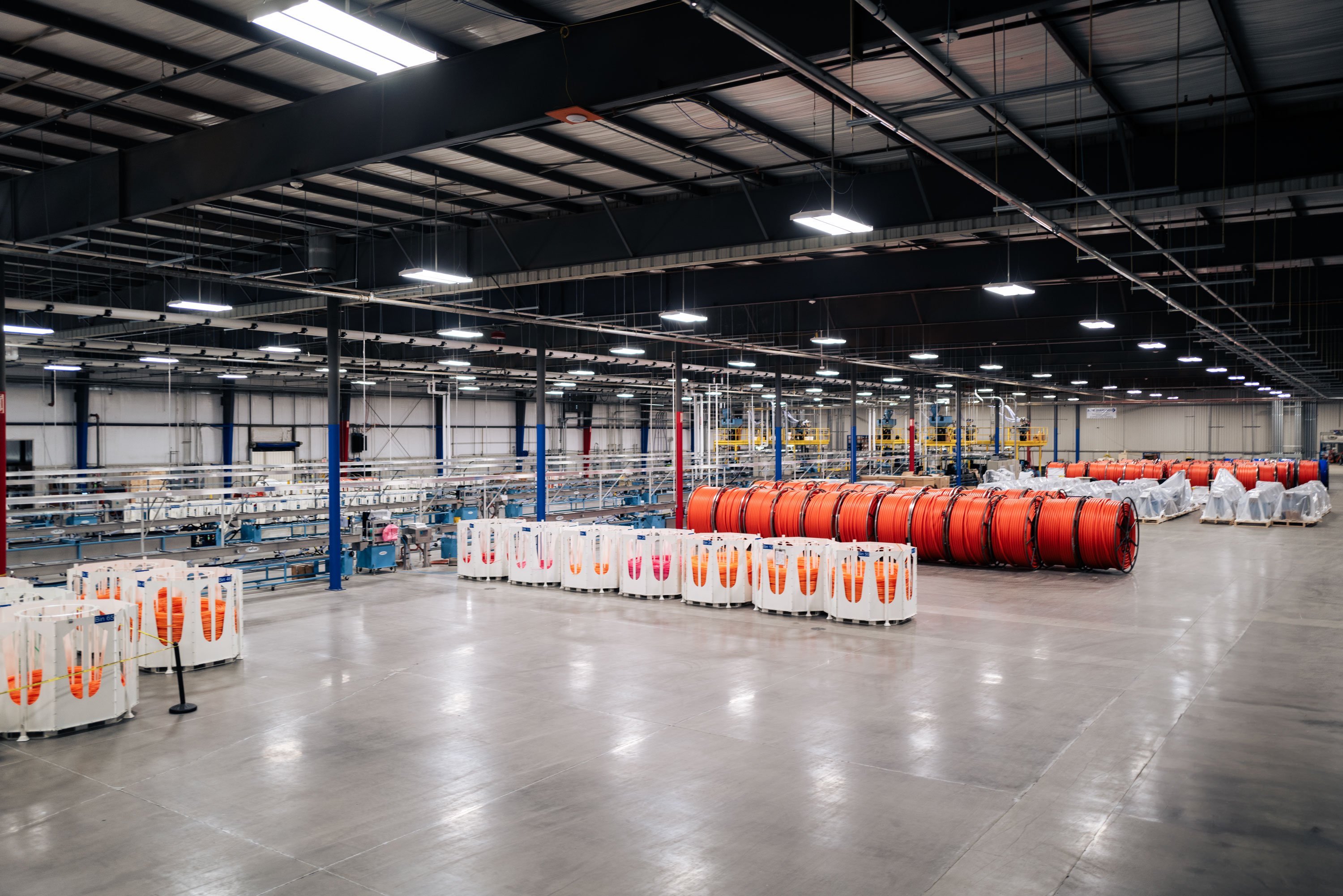 This is a well-lit warehouse with rows of large spools with orange micro duct. There are no people visible in this image.