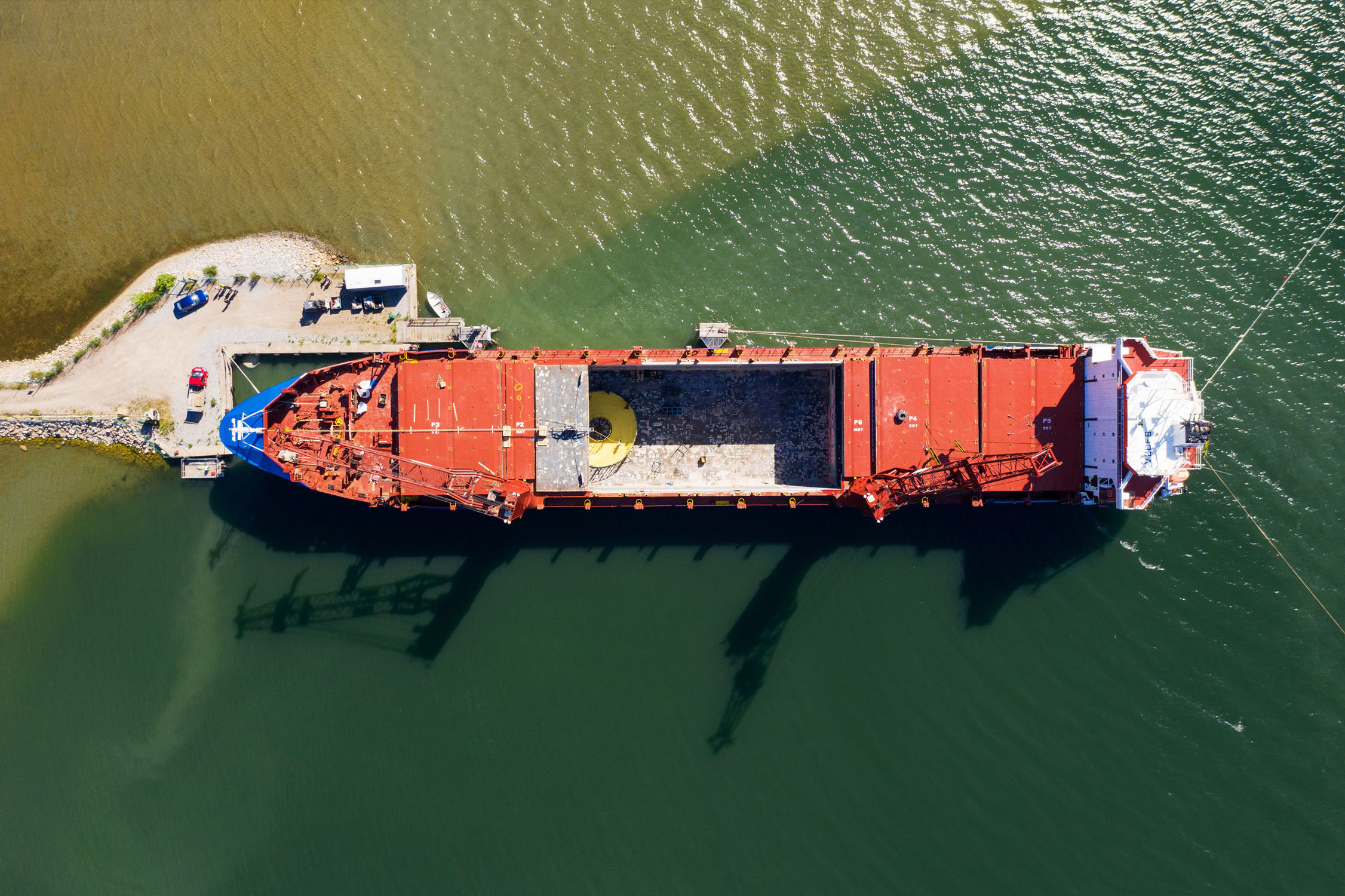 Aerial view of a large red cable vessel docked at a pier. The water surrounding the ship is greenish in color, indicating it might be in a harbor.