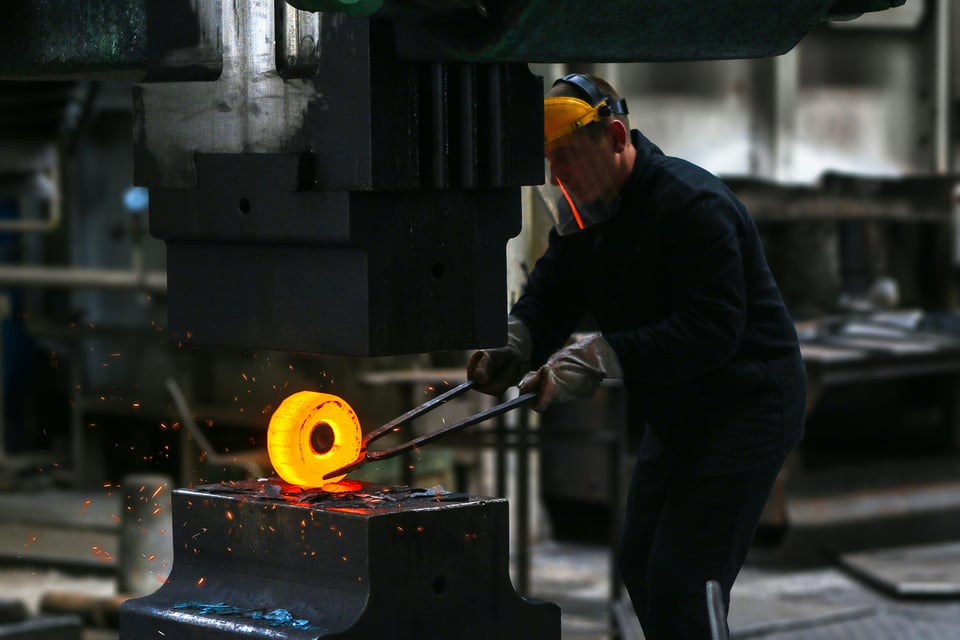 A person in a workshop using tools to shape a circular piece of glowing metal under a large press. Sparks are flying as the metal is being worked on.
