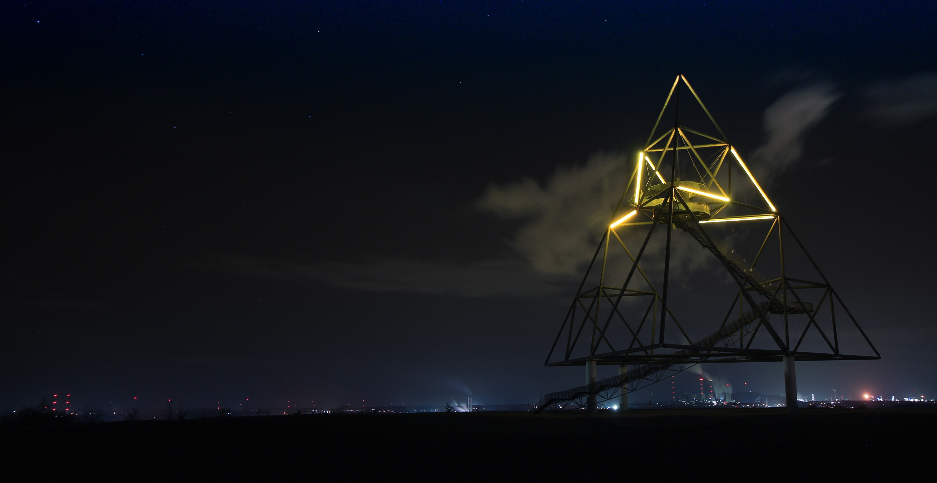 A metallic, star-shaped structure illuminated against a dark night sky. Distant city lights and stars are visible in the background.
