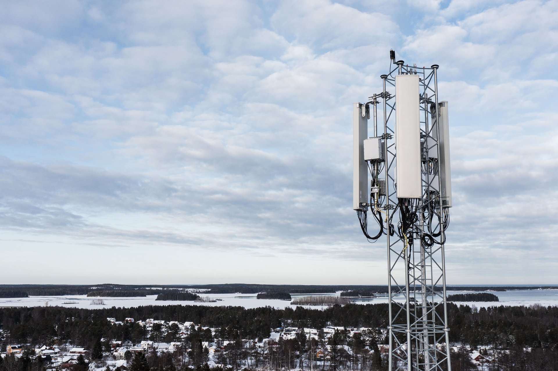 This is a tall cell tower with antennas and equipment. It stands in a snowy landscape with scattered buildings and water bodies.