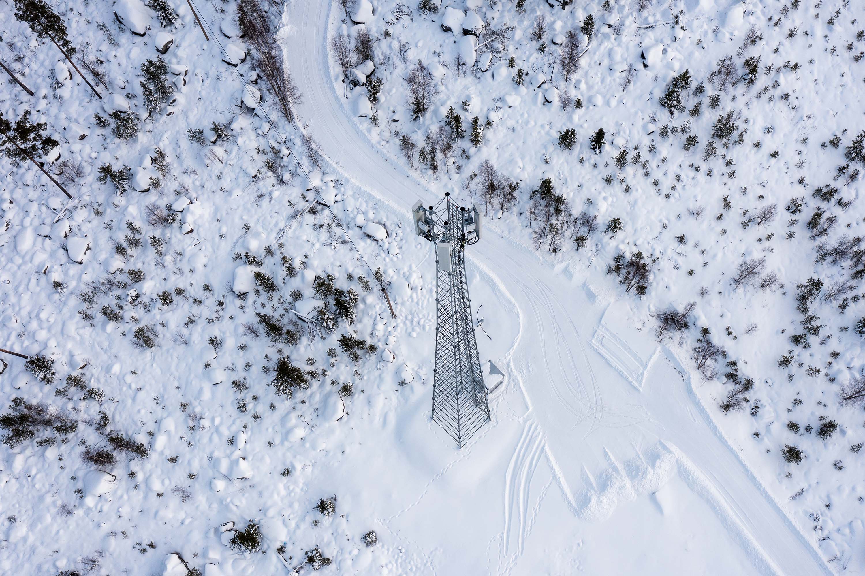 This is an aerial view of a snowy landscape with a metal 5G tower in the center. The ground is covered in snow, with a curved path on the left side.