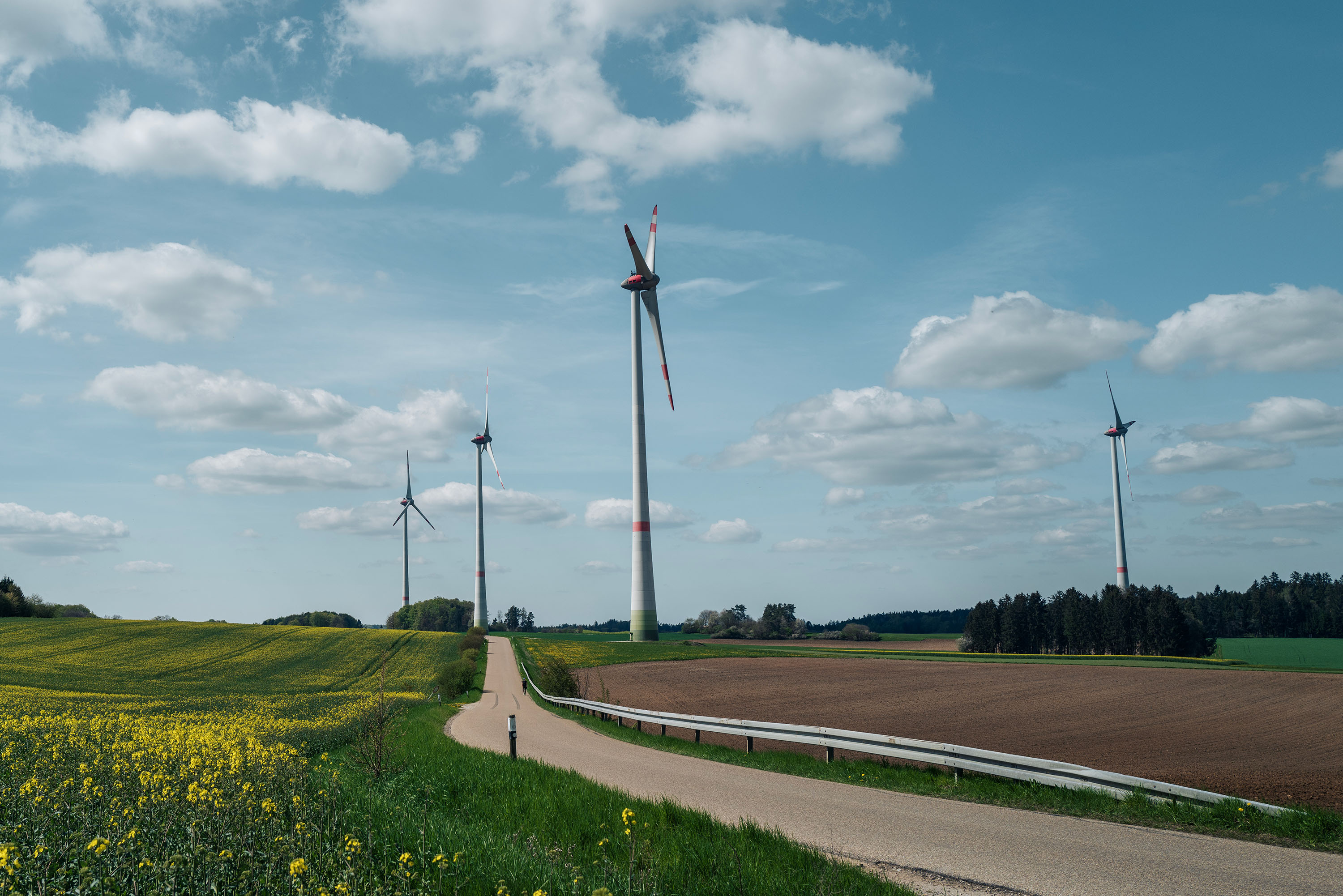 A rural area with four wind turbines standing tall on a green field next to a country road. The partly cloudy sky adds depth to the scene.