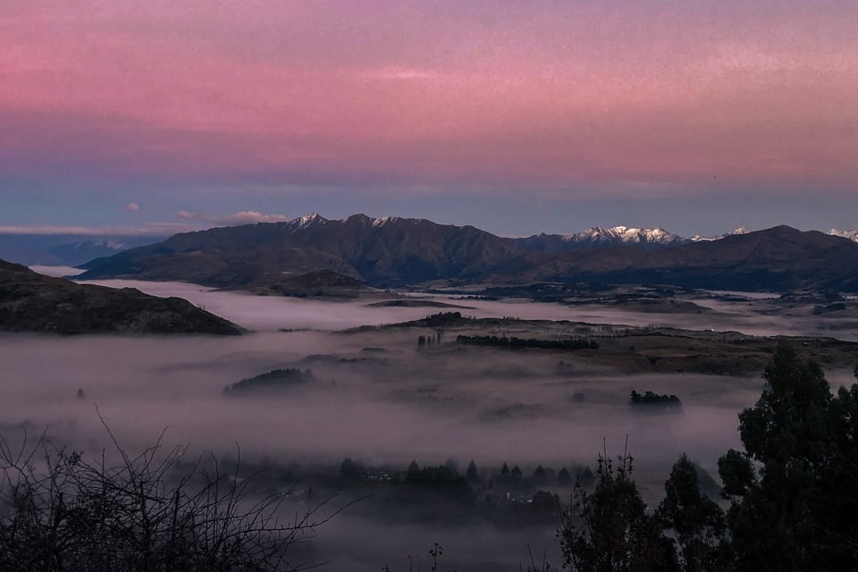 A view of a landscape with mountains and trees at dusk. The fog lies like a lake over the ground.