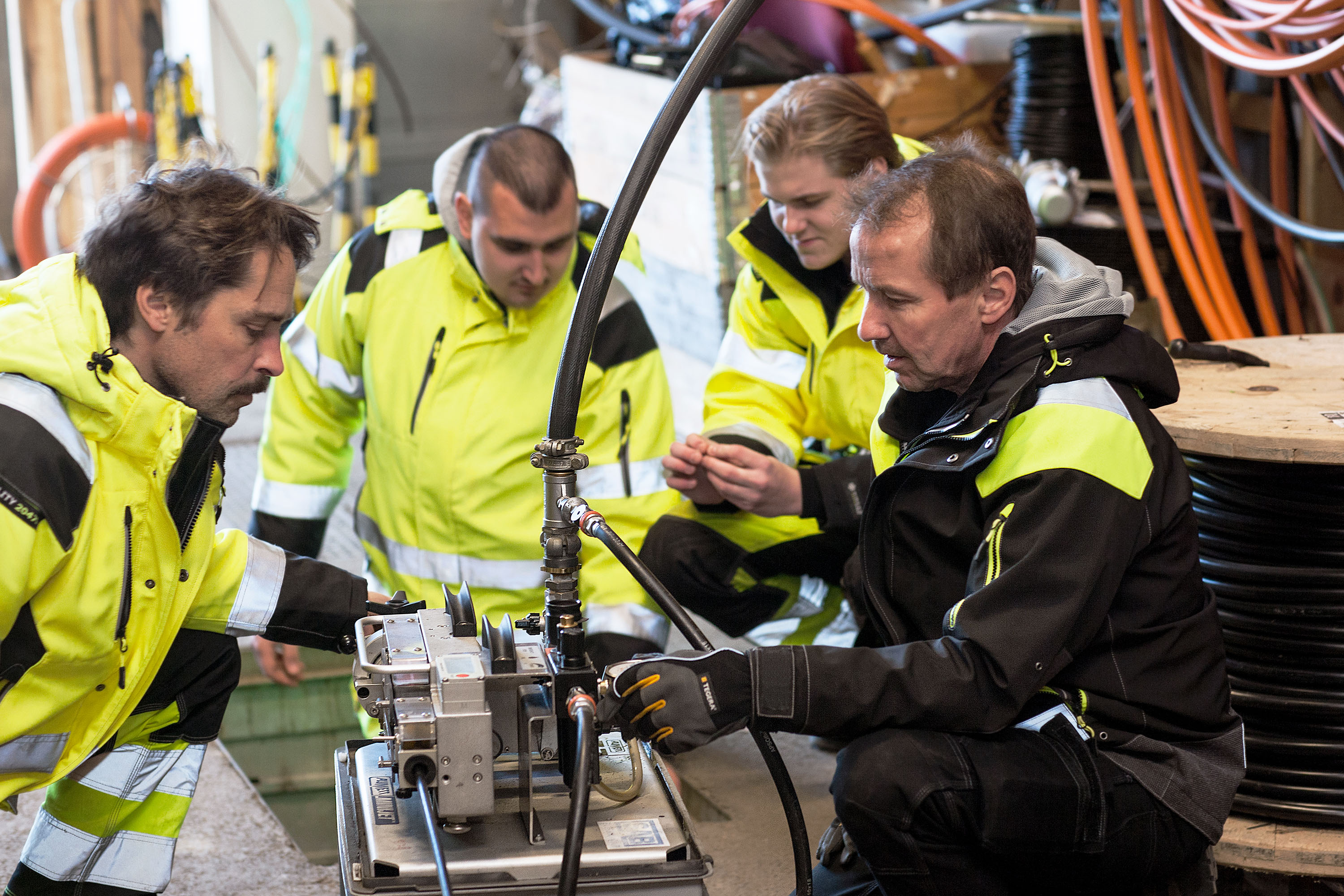 A group of four people in safety jackets, working on a mechanical device indoors. They seem to be collaborating on a repair or training session.