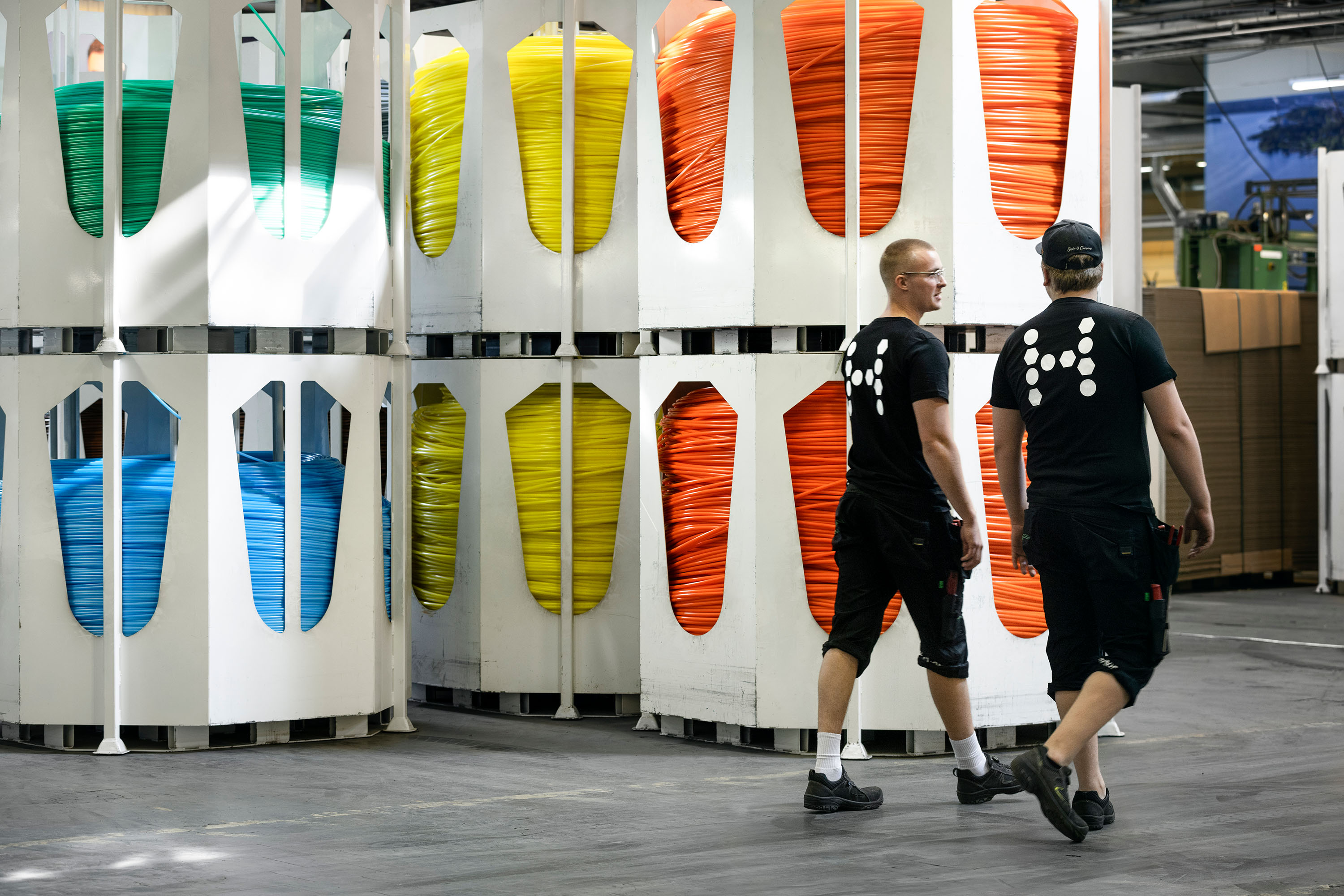A warehouse with large spools of micro ducts in different colors. Two workers with black t-shirts with the Hexatronic logo on the back are walking by.