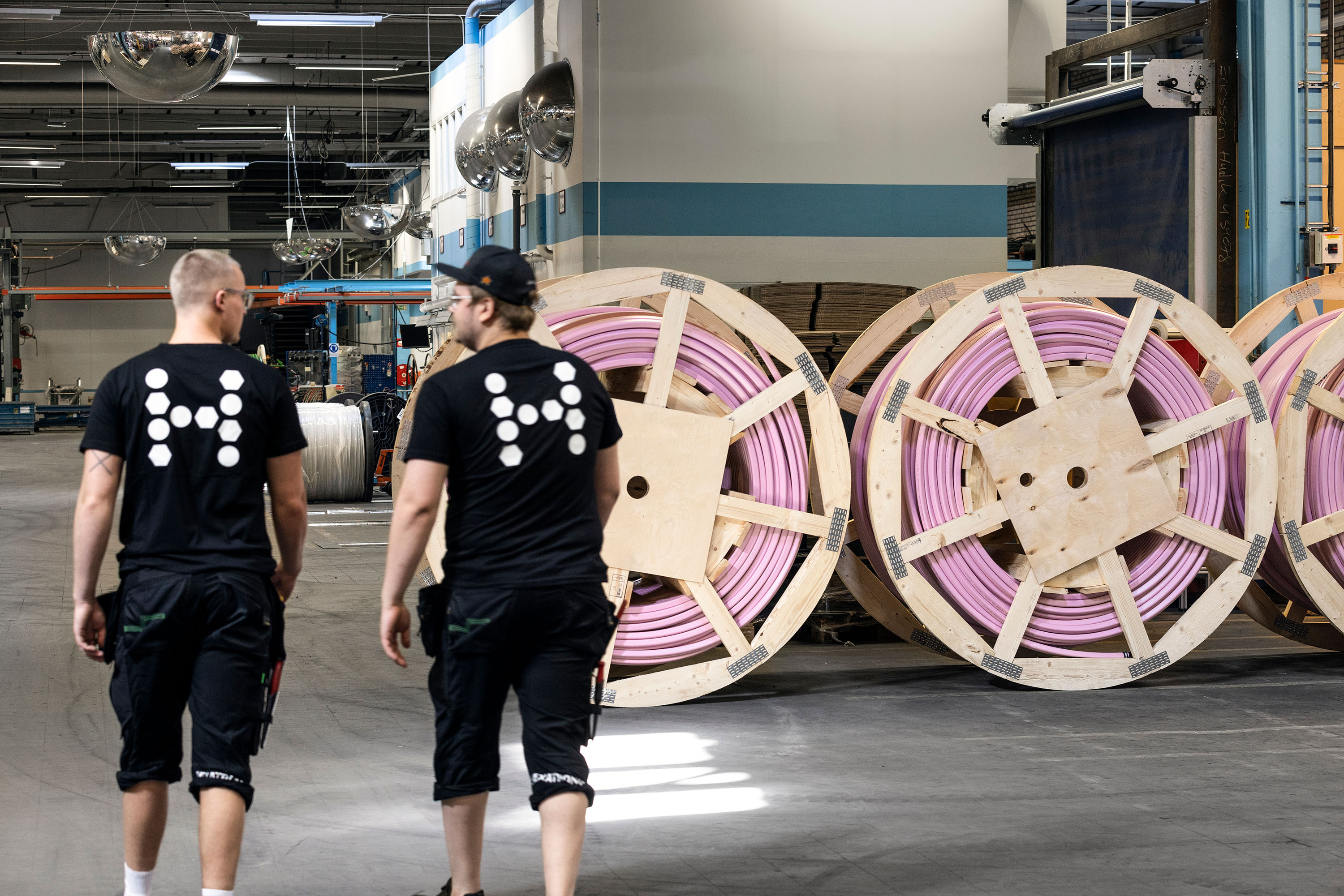 Two people in an industrial space with high ceilings and bright lighting. They are walking past giant spools of pink micro duct.