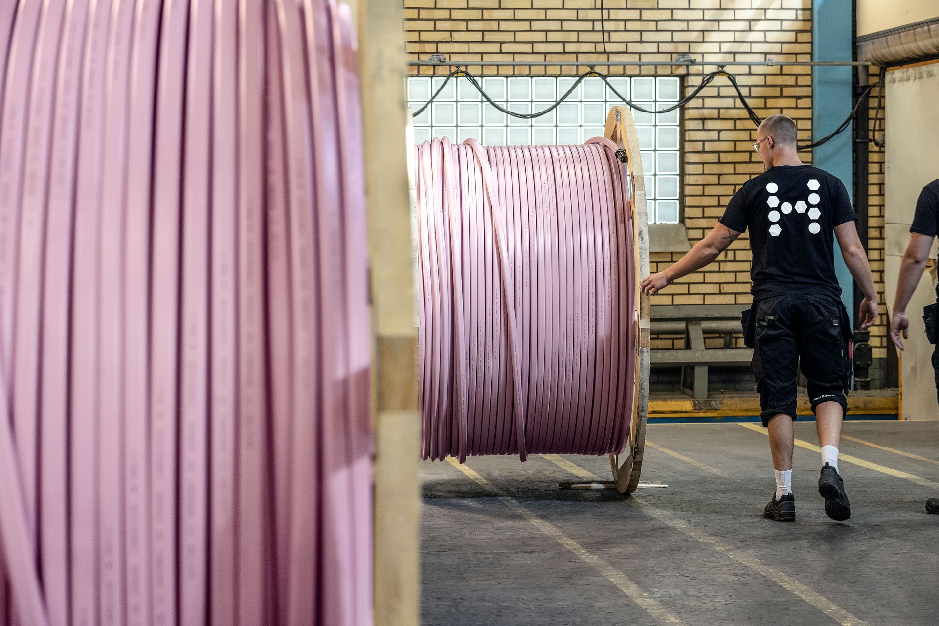 A person in a warehouse walks past a large spool of pink microduct. More coils and industrial elements are visible.