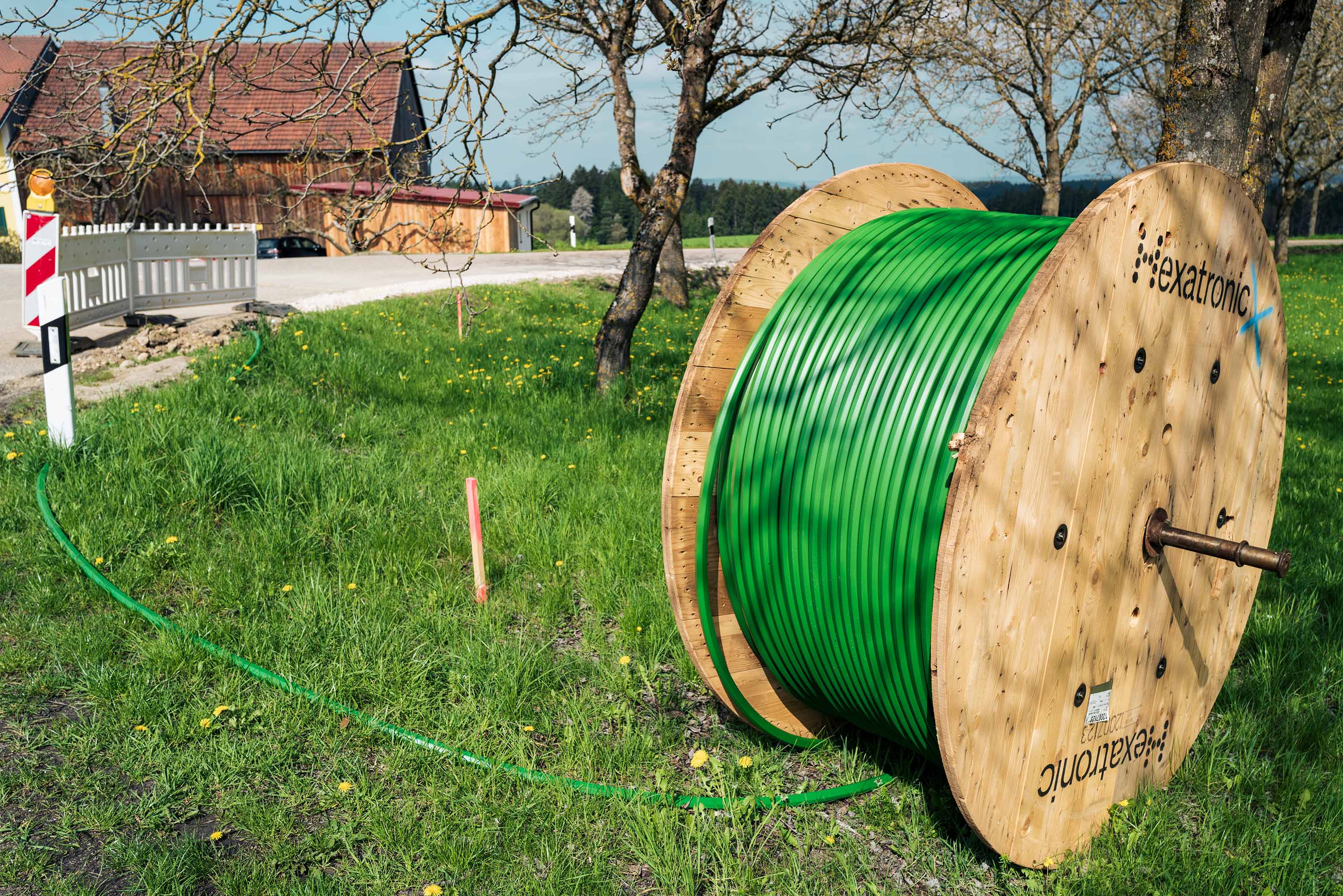 A large wooden spool with green duct is by a grassy roadside, near a house and fence, under a clear sky.