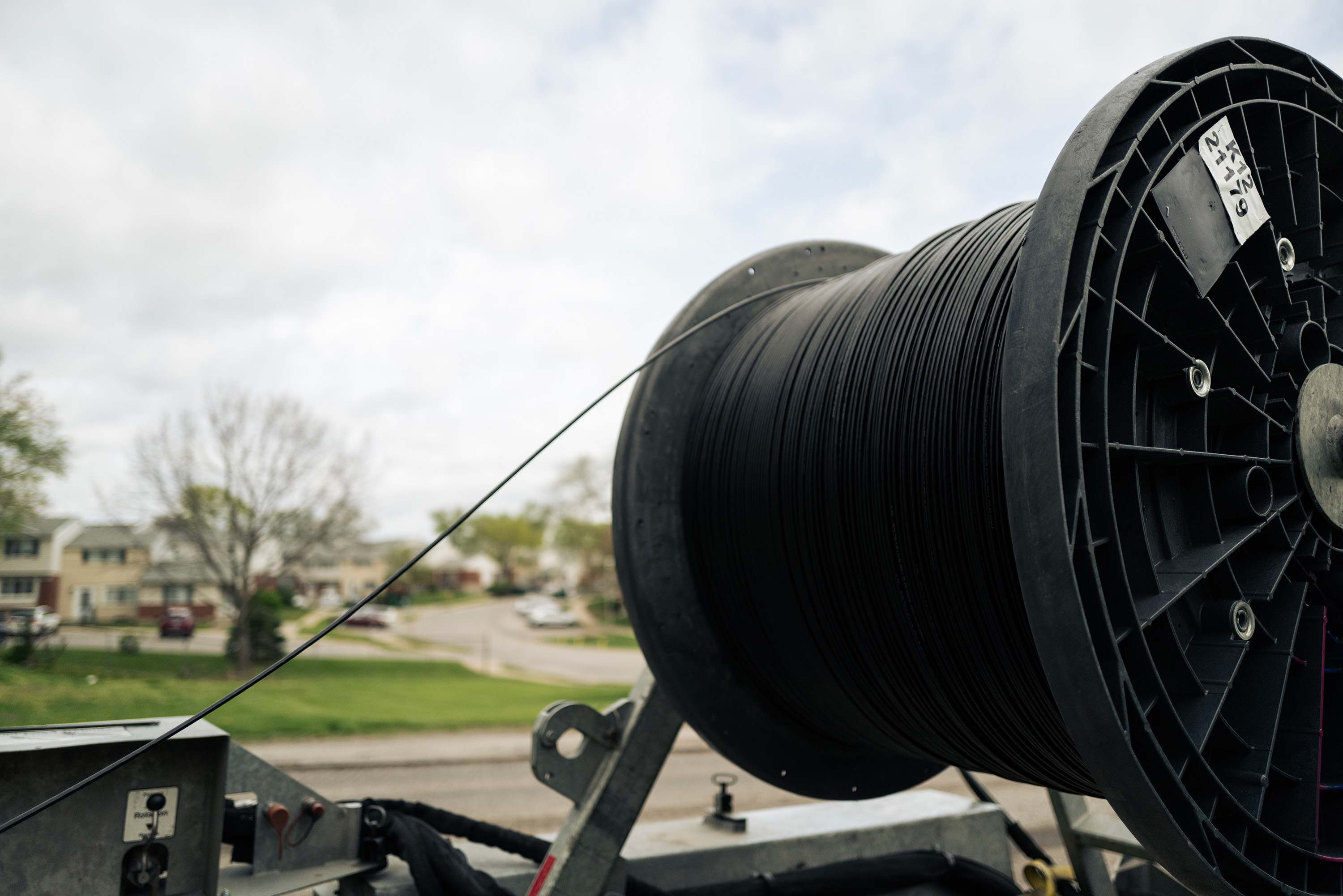 A large spool of black fiber optic cable on a trailer in a suburban neighborhood under a cloudy sky, suggesting ongoing work.