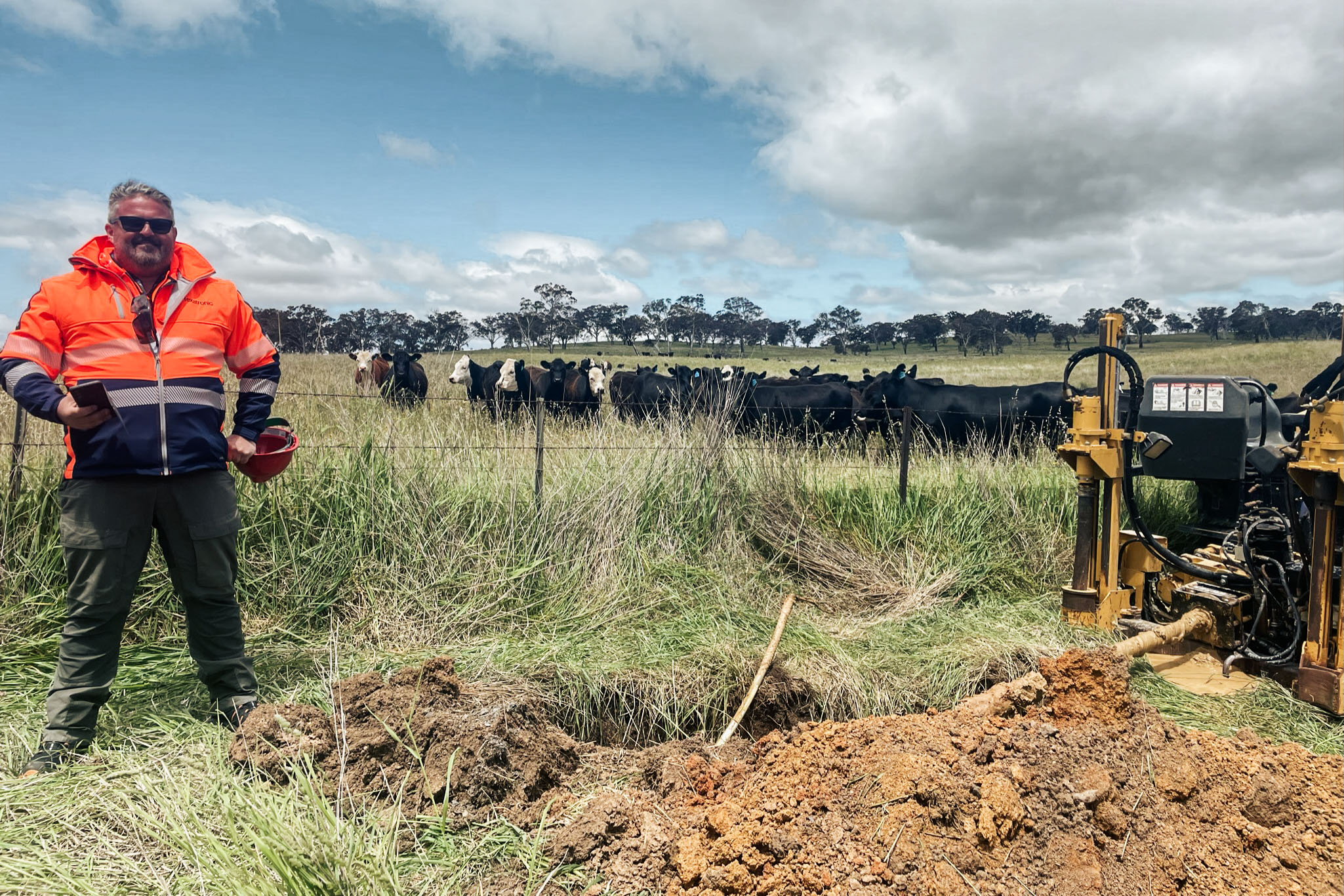 A worker in safety gear stands by machinery in a field. Cows graze under a cloudy sky in the distance.