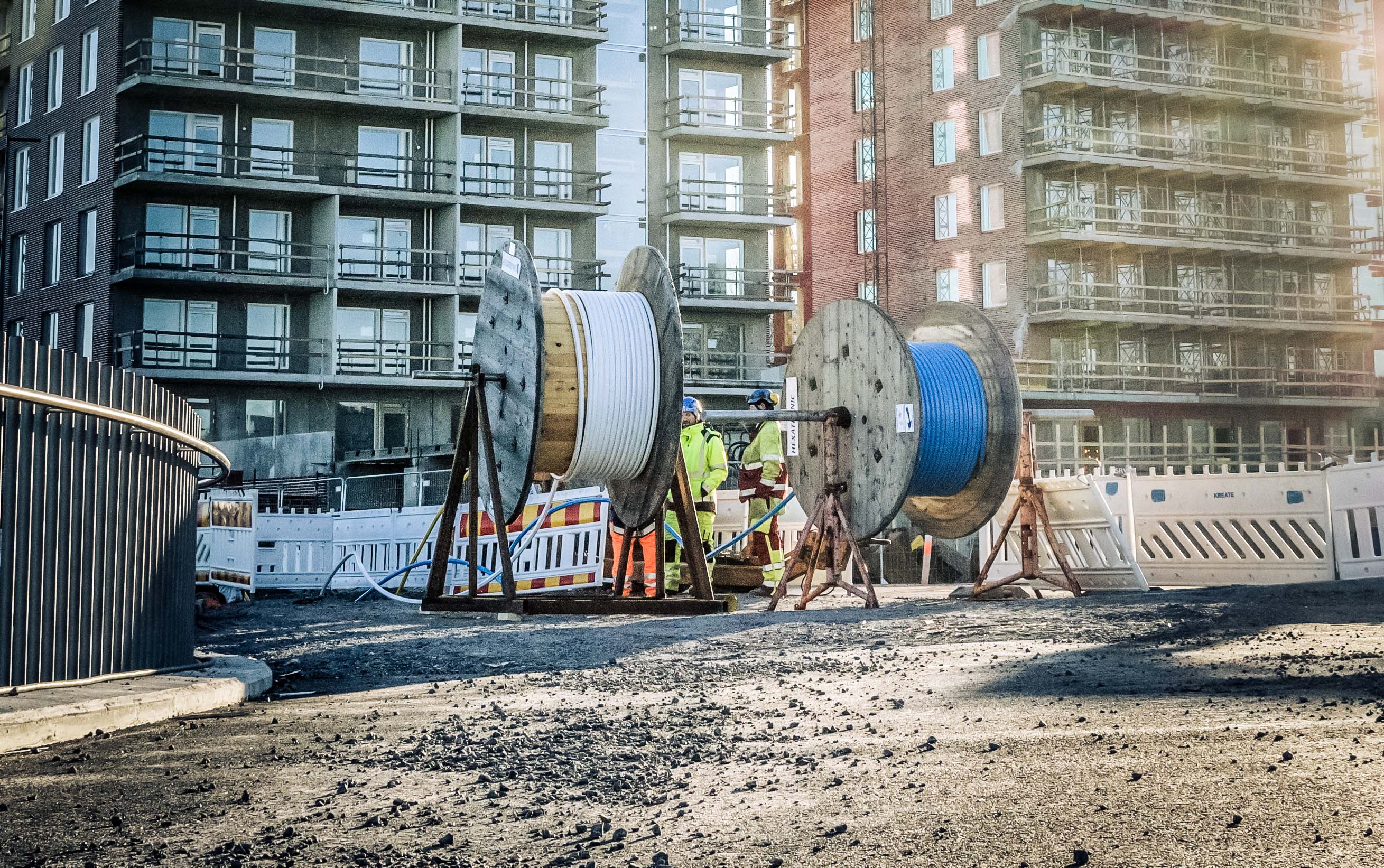 People in safety clothing and protective helmets work on a construction site. In the foreground are large wooden drums with microduct.