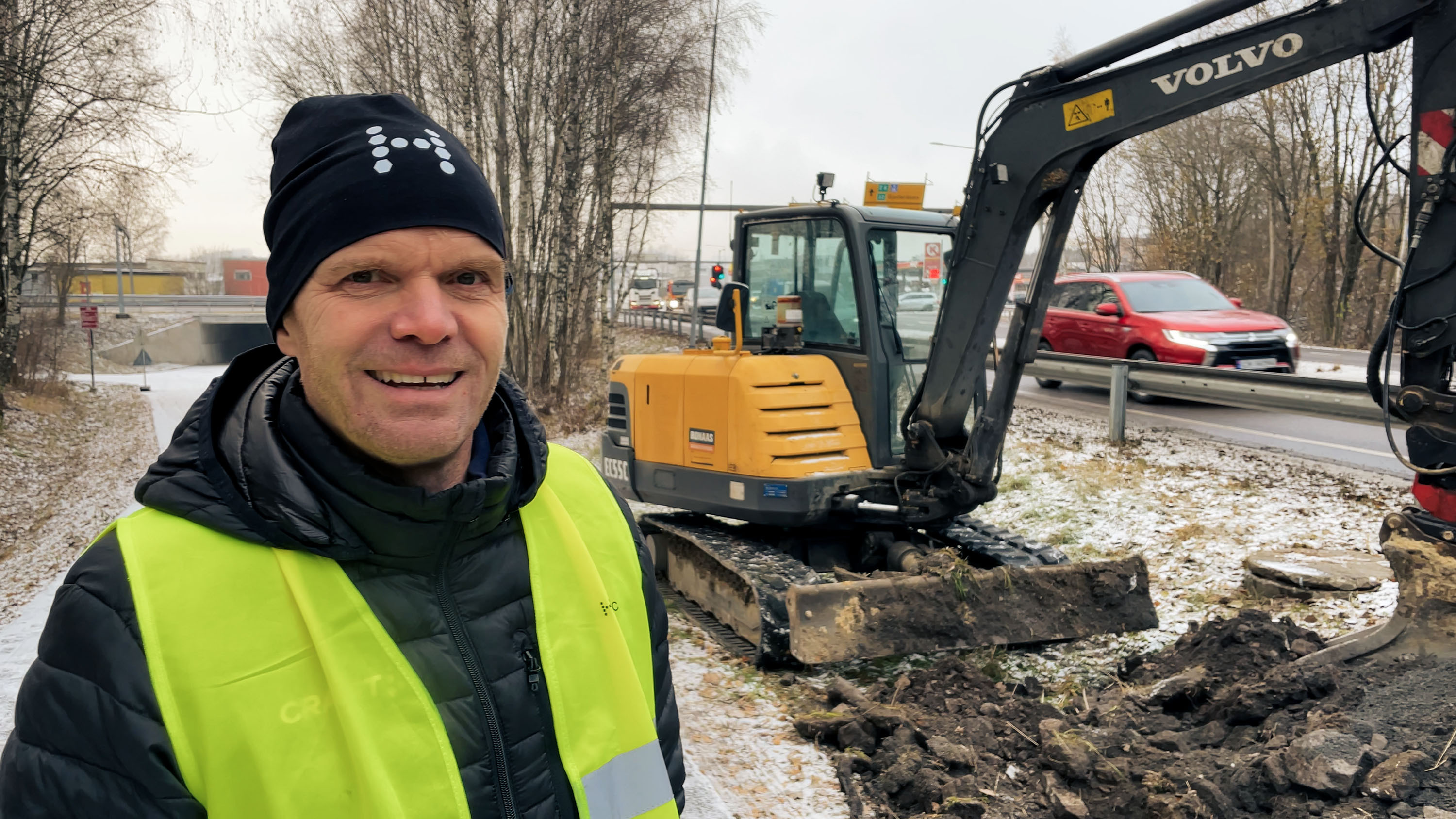 A person from Hexatronic Field Support in a high-visibility vest stands near a Volvo excavator on a snowy, muddy construction site in Norway. 