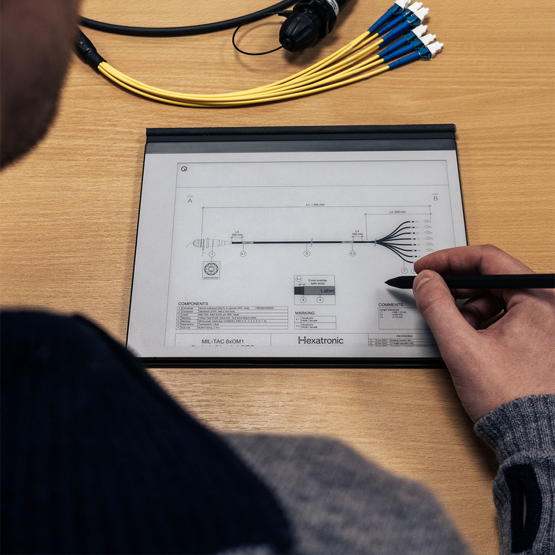 A person is studying a technical drawing on a wooden table, with cables and connectors nearby. They’re holding a pen, possibly making notes.