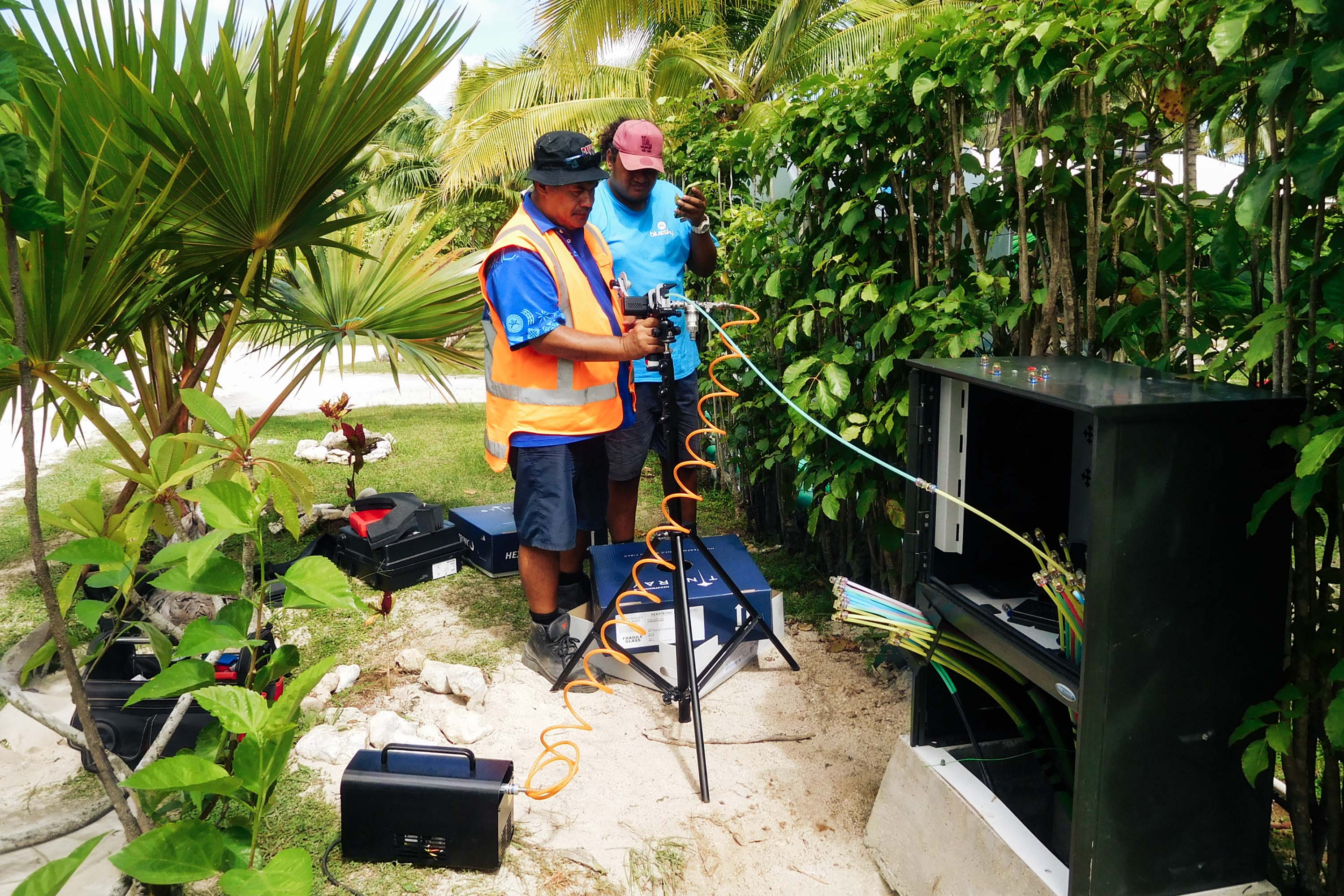 Two people install Stingray Air Blown Fiber into a cabinet. The surroundings are green, with palm trees and sand.