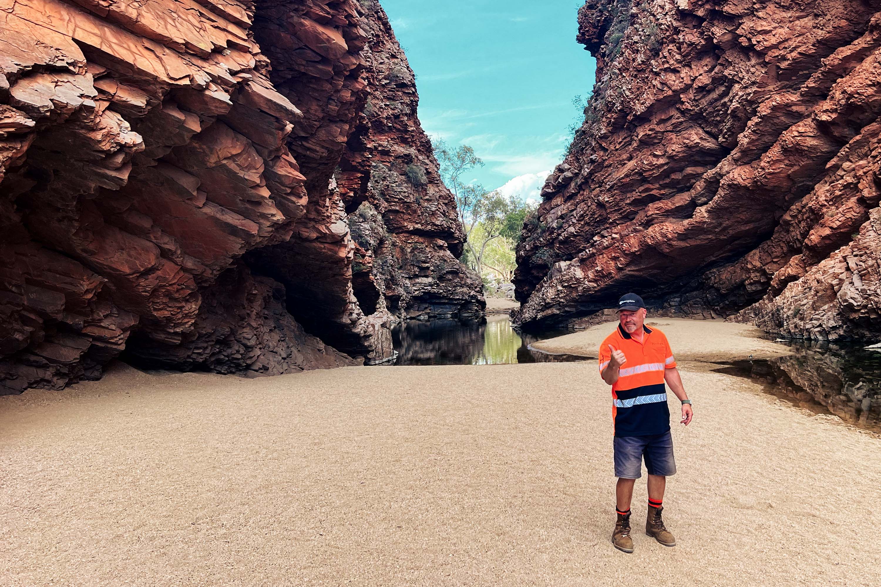 A person in orange work clothes stands on a beach between two rocks and gives the thumbs up.