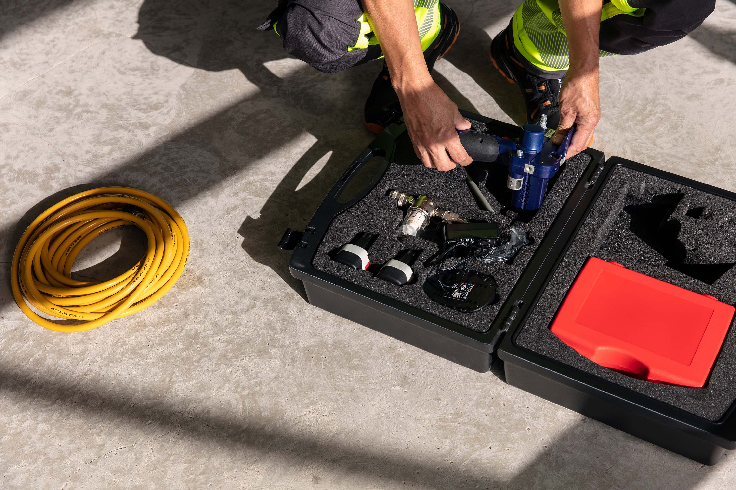 A person in safety gear is kneeling on a concrete floor, organizing a toolbox with various tools, next to a coiled yellow hose.