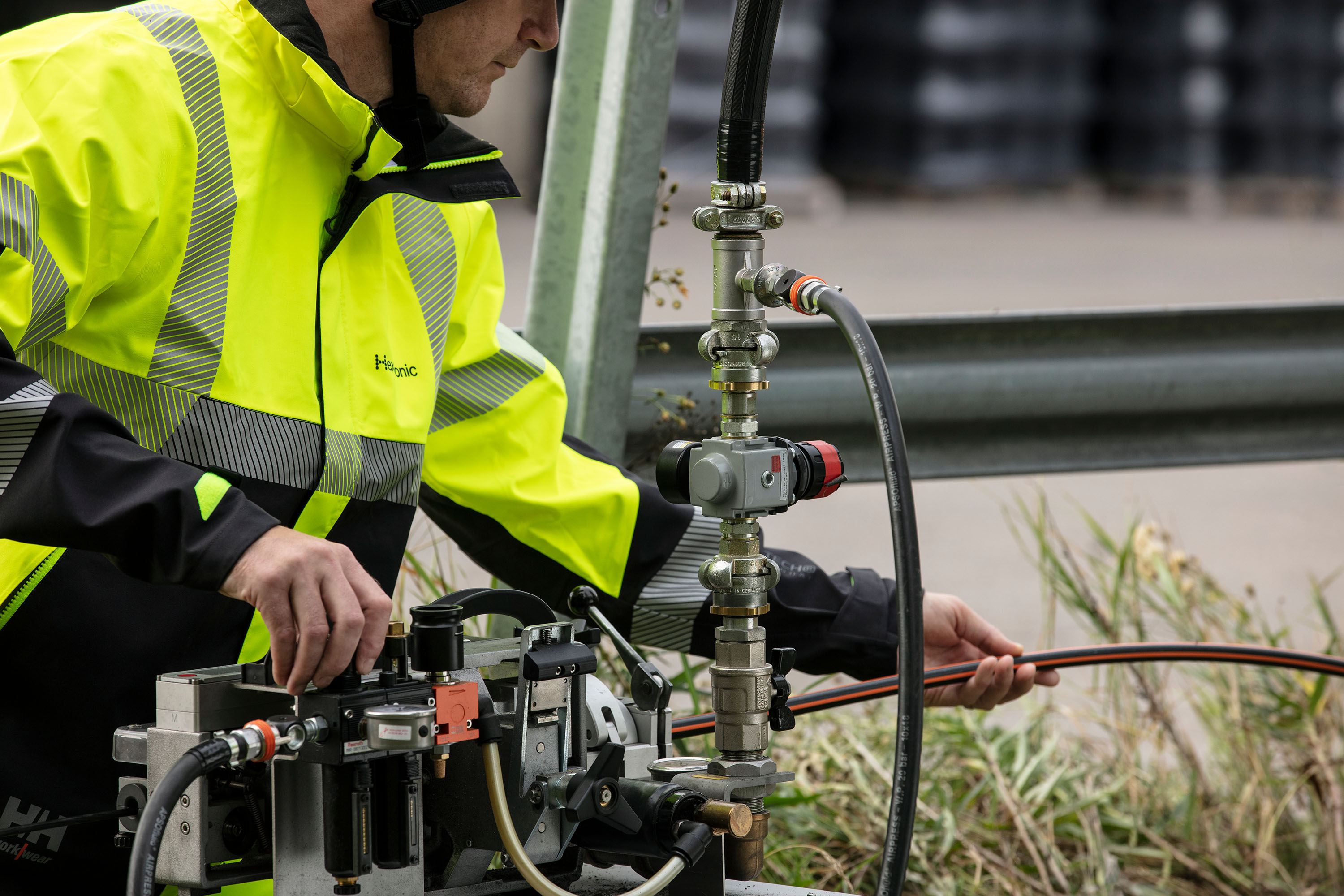 A person in a high-visibility jacket is outdoors, operating a fiber optic installation tool connected to a duct. It appears to be an industrial site.