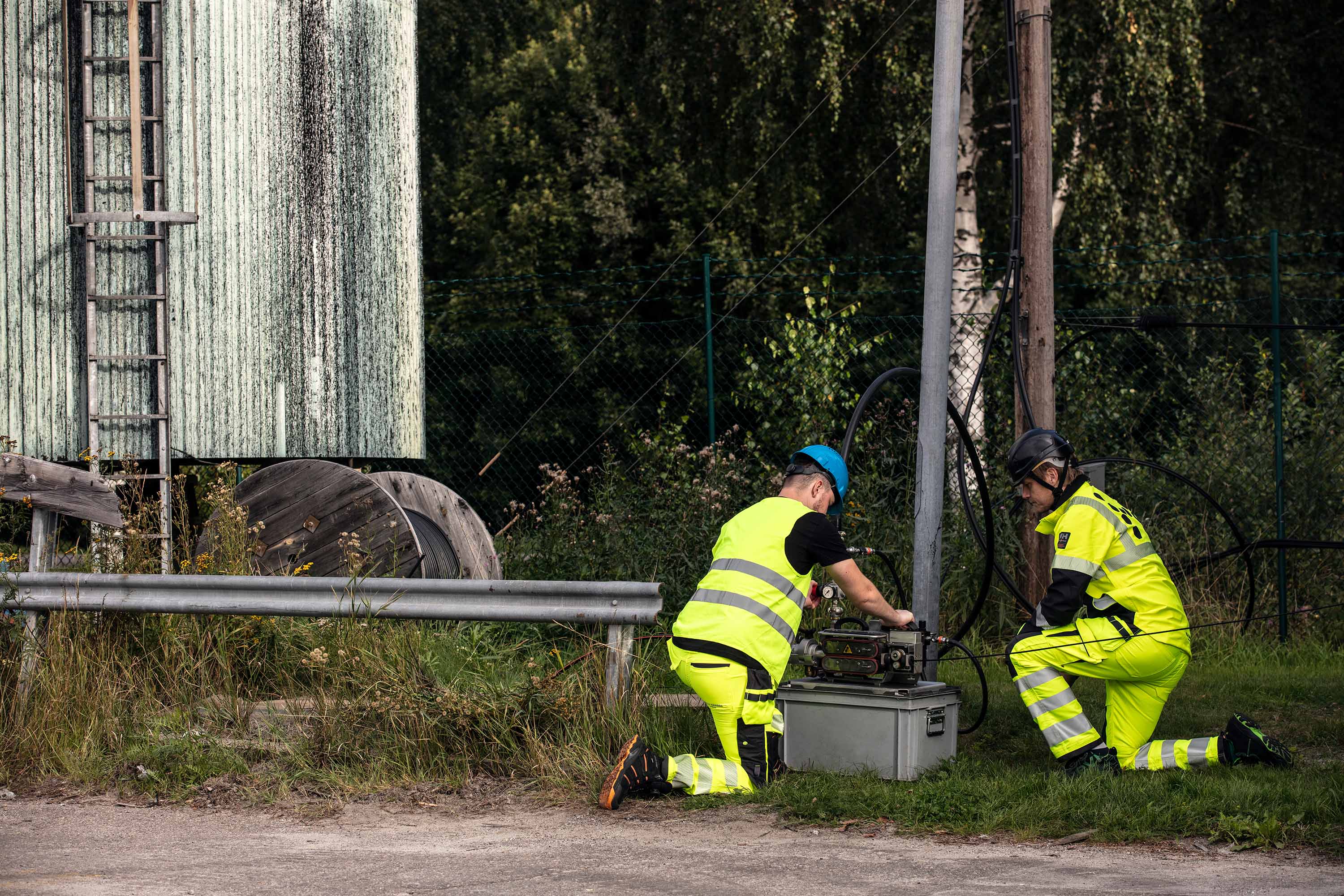 Two workers in yellow safety gear are attending to equipment near a large green structure, surrounded by greenery and a metal fence.