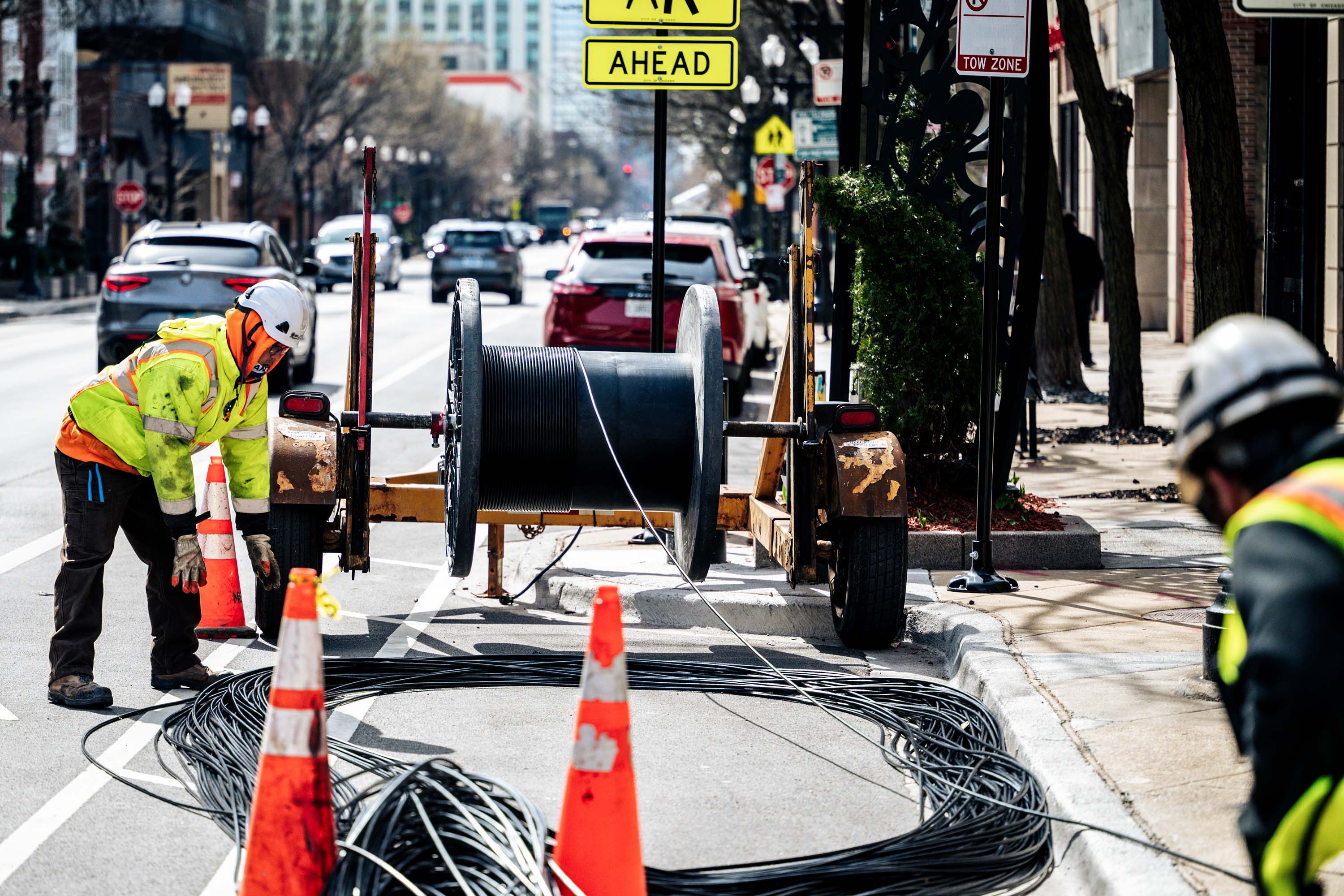 Two workers in bright safety gear is handling cables on a city street, surrounded by traffic cones for safety.