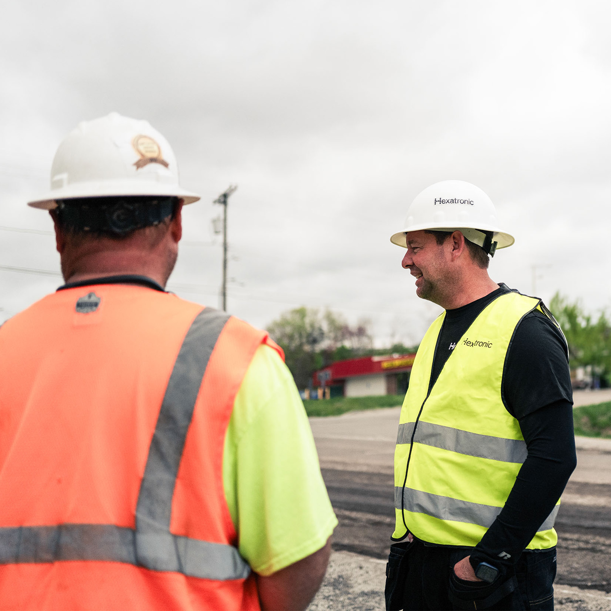 Two workers in safety gear stand near a road, possibly discussing a project. A traffic cone indicates a work area.