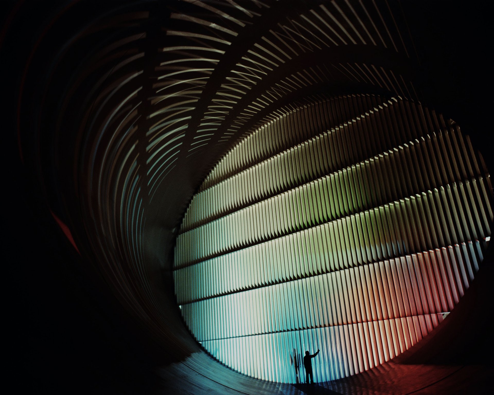 A person stands in a large, ribbed tunnel bathed in soft, gradient lighting from green to blue to red, creating a serene atmosphere.