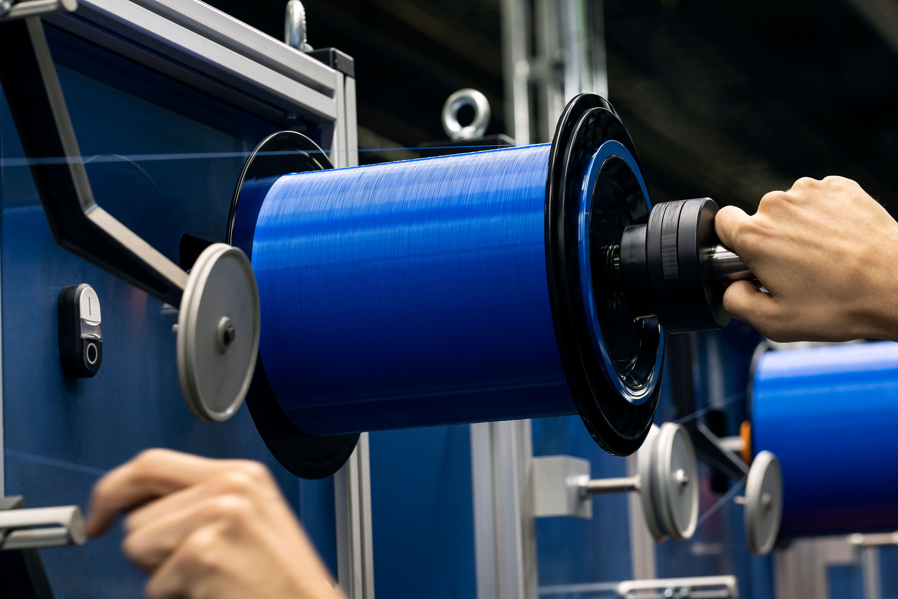 Hands attach a black spool of blue fiber thread to a machine in a factory setting.
