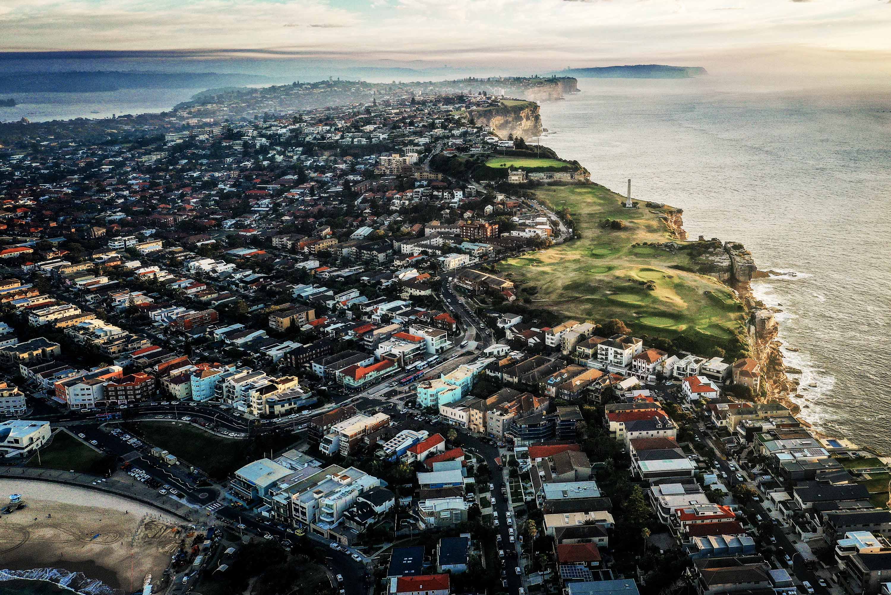 Aerial view of a coastal town at dusk, dense buildings, green cliffside, vast ocean extending into the horizon.