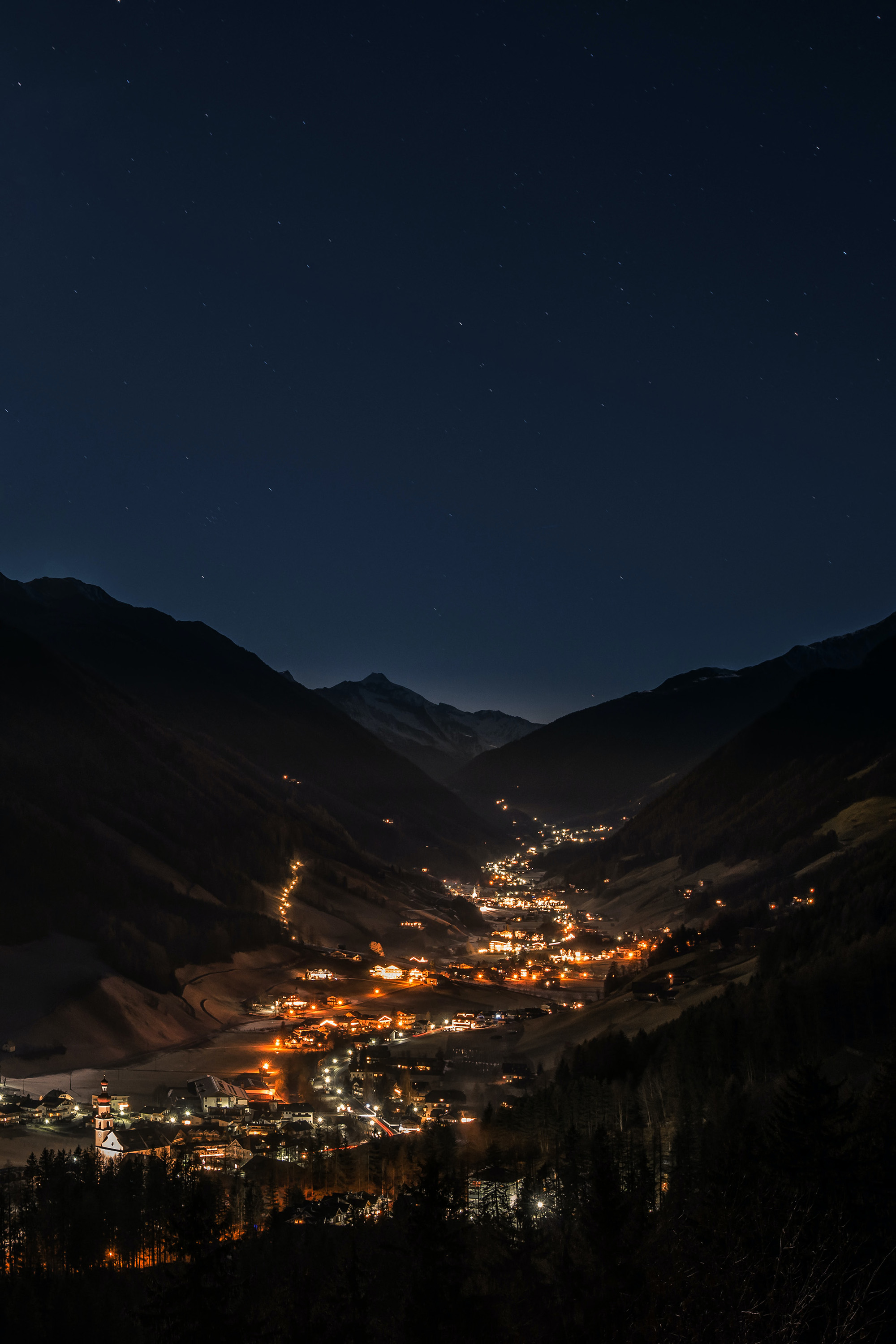 A tranquil night in a mountain valley. A town’s warm lights glow, surrounded by dark mountains under a starry sky.